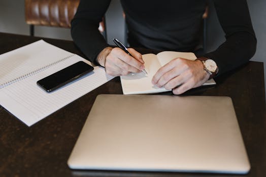 A person writing in a notebook with a laptop and smartphone on a table.